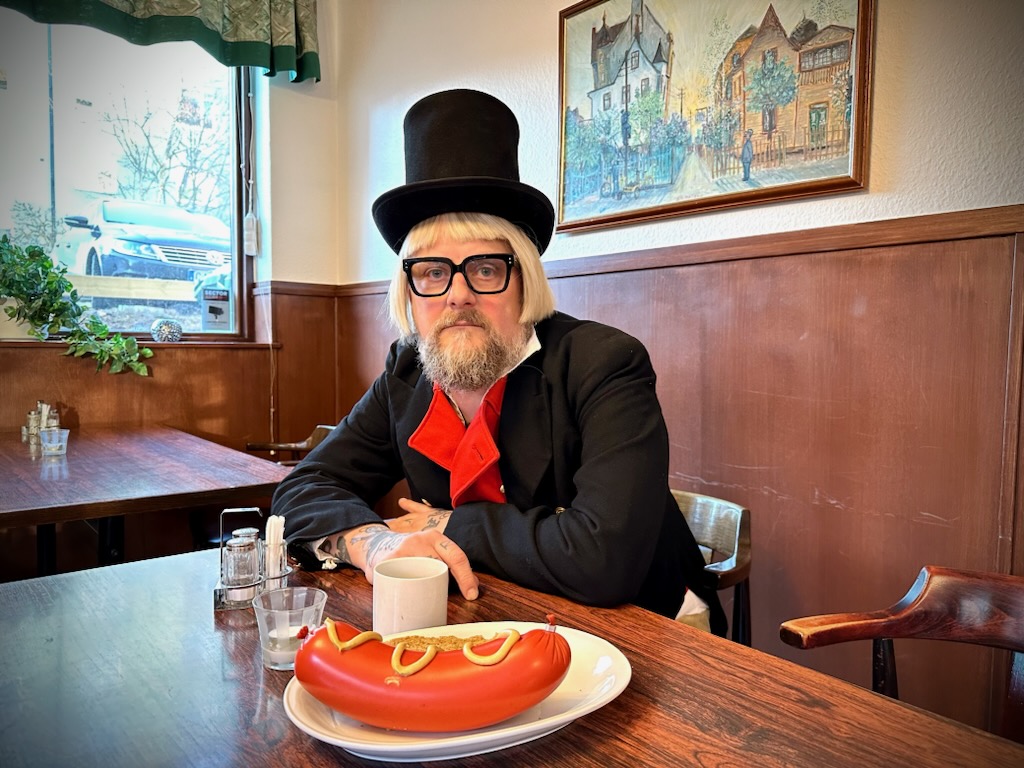 Bild på Patrik Arve i hög hatt och med en falukorv framför sig på bordet