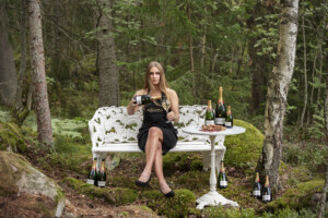 Porträttbild på Sara Bernhardsson på parkbänk i skogen med champagne Bollinger